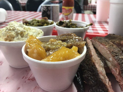 A plate of barbecue brisket with sides of peach cobbler, potato salad, and greens on a checkered tablecloth.