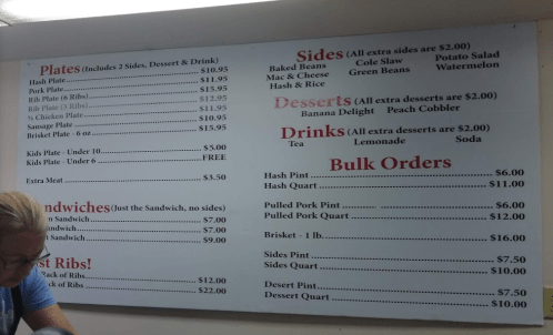 Menu board displaying prices for plates, sides, desserts, drinks, sandwiches, and bulk orders at a restaurant.