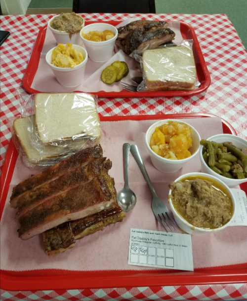 Two red trays with barbecue ribs, pulled pork, sides of green beans, peach cobbler, and bread on a checkered tablecloth.
