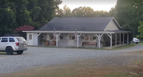 A small, one-story building with a porch, surrounded by trees and gravel parking, in a rural setting.