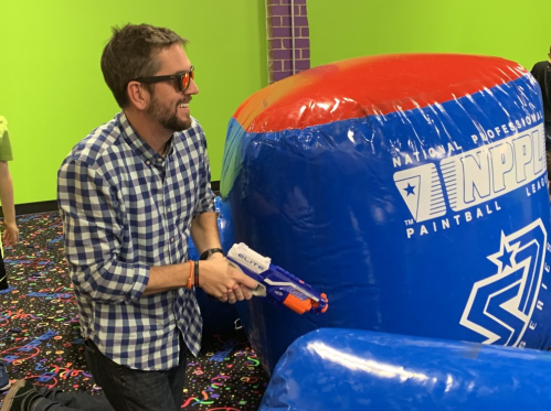 A man in sunglasses playfully aims a toy gun at a colorful inflatable structure in a vibrant indoor setting.