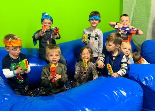 A group of eight children in colorful bandanas, holding toy guns, smiling in a blue play area with a green background.