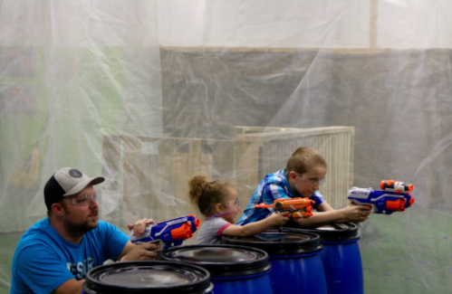 Two children and an adult aim toy blasters from behind blue barrels in a playful indoor setting.