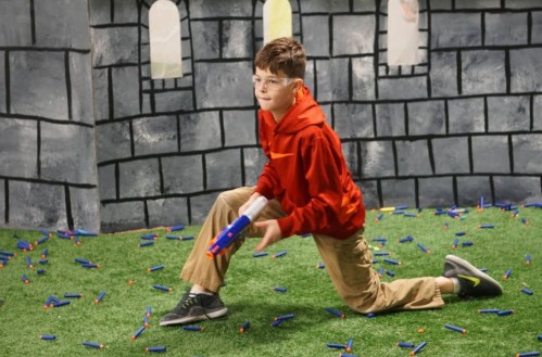 A boy in a red hoodie kneels on a grassy surface, aiming a toy blaster in a castle-themed play area filled with darts.
