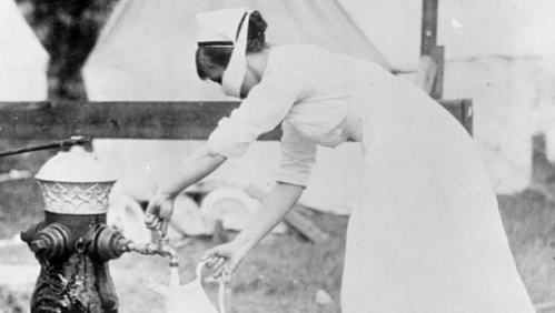 A nurse in a white dress fills a pitcher from a fire hydrant in a historical black-and-white photo.
