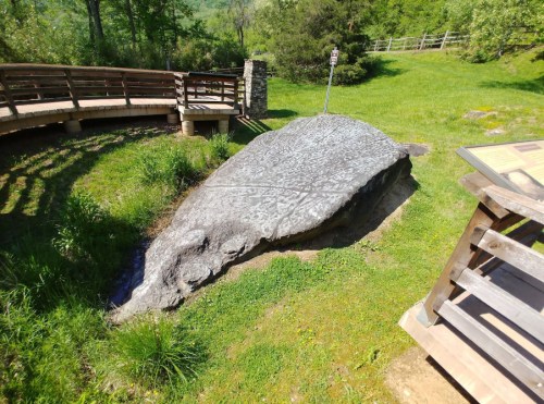 Large rock with ancient carvings, surrounded by grass and trees, near a wooden viewing platform and informational sign.