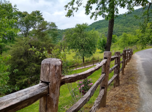A rustic wooden fence lines a scenic road, surrounded by lush greenery and rolling hills under a cloudy sky.
