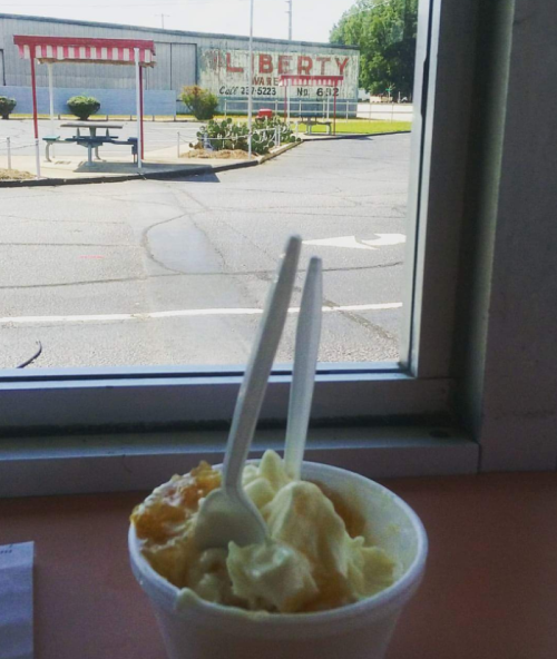 A cup of ice cream with two spoons sits on a table, with a view of a building and street outside the window.
