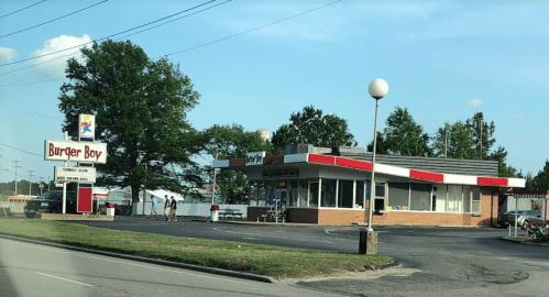 A fast-food restaurant named "Burger Boy" with a drive-thru and outdoor seating, surrounded by trees and a clear sky.
