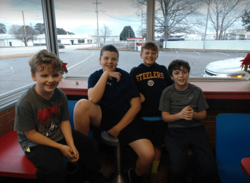 Four boys sitting together at a table in a casual setting, smiling and enjoying each other's company.