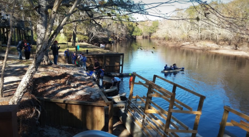 A group of people near a river, with some in kayaks and others on the shore by a wooden dock and trees.