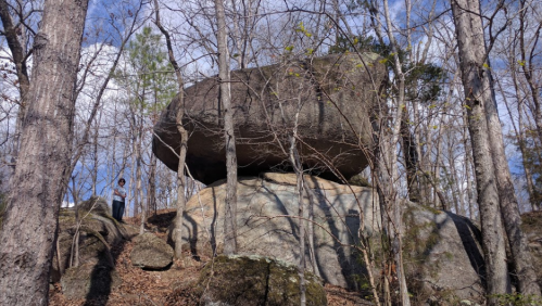 A large boulder balanced on a rock formation, surrounded by bare trees and a blue sky with scattered clouds.