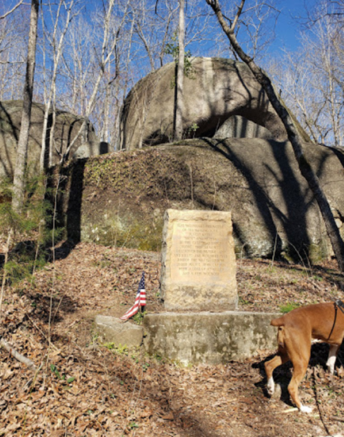 A stone monument with an American flag stands near large boulders in a wooded area under a clear blue sky.