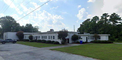 A white, single-story building surrounded by green grass and trees, with a gravel driveway and blue recycling bin.