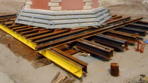 Construction site featuring steel beams and supports under a structure, with a yellow beam and a barrel visible.