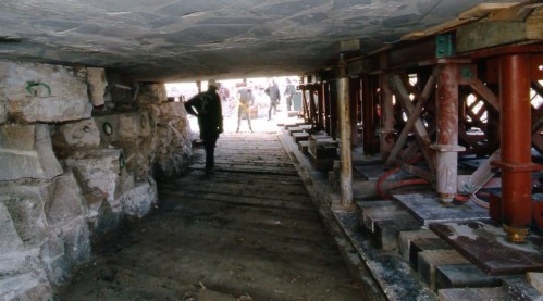 A person walks under a structure supported by metal beams and wooden planks, with workers visible in the background.