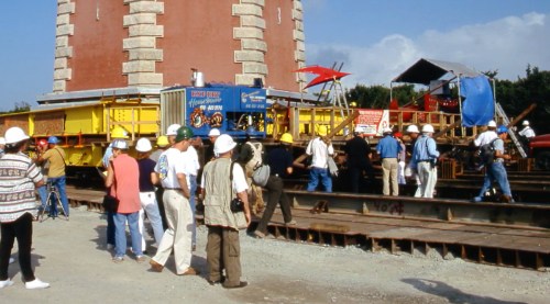 A group of people in hard hats gathers around a train car near a tower, with equipment and tents in the background.