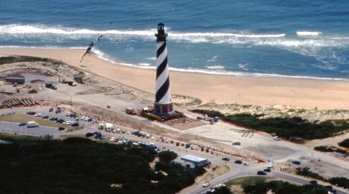 Aerial view of a striped lighthouse near a sandy beach, with waves crashing and vehicles parked nearby.