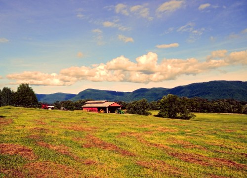 A scenic landscape featuring a barn in a green field, surrounded by mountains and a blue sky with fluffy clouds.