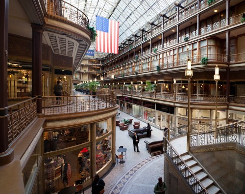 Interior view of a historic atrium with shops, seating areas, and a large American flag hanging above.