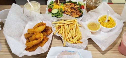 A table spread with fried onion rings, French fries, a salad, mac and cheese, and drinks in clear cups.