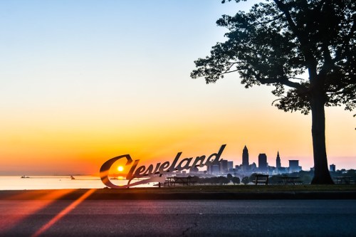 Sunrise over Cleveland skyline with a large "Cleveland" sign in the foreground, framed by trees and water.