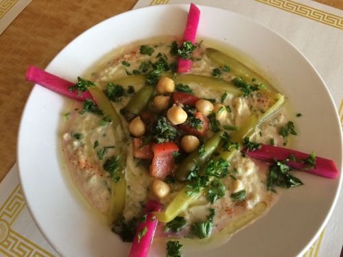 A plate of creamy dip topped with chickpeas, diced vegetables, and garnished with herbs and colorful pickled vegetables.