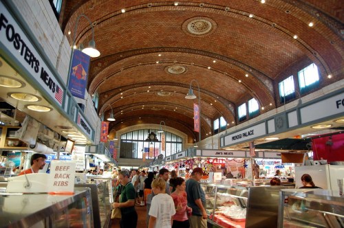 A bustling market interior with high brick arches, vendors, and shoppers exploring various food stalls.