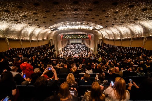A crowded theater with a large choir on stage, ornate ceiling, and audience members taking photos.
