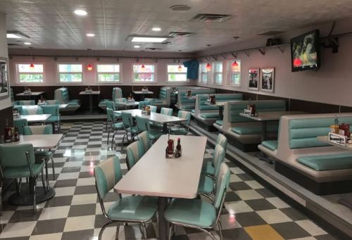 A retro diner interior with teal booths, checkered floor, and tables, featuring vintage decor and a TV on the wall.