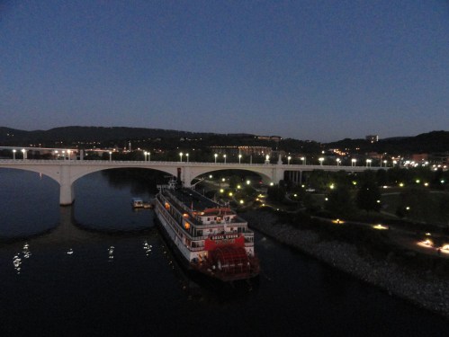 A riverboat docked near a lit bridge at dusk, with city lights reflecting on the water.