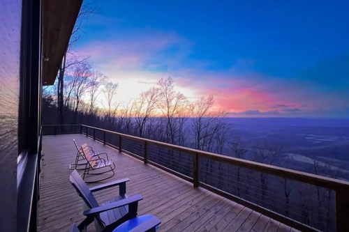 A wooden deck with chairs overlooks a colorful sunset and a valley surrounded by trees.