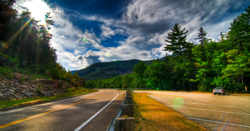 A scenic road curves through lush greenery under a dramatic sky, with mountains in the background and a parking area nearby.