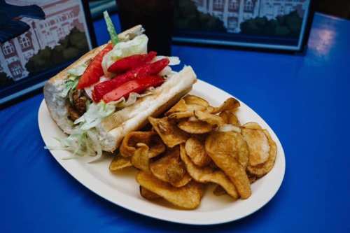 A sandwich with lettuce, tomatoes, and pickles served with a side of crispy potato chips on a white plate.