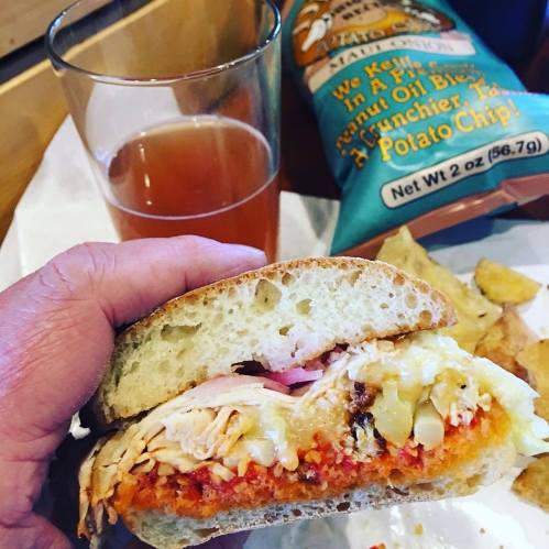 A hand holding a sandwich filled with chicken, cheese, and red sauce, alongside a glass of beer and a bag of chips.