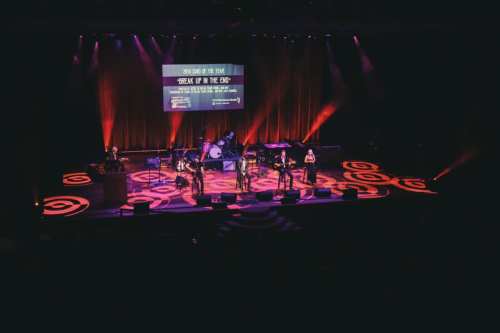 A live band performs on stage with colorful lights and a large screen in the background.