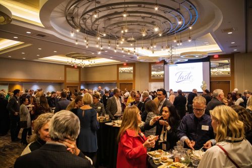 A crowded event space with people mingling, enjoying food, and a large screen displaying "Welcome to Taste."