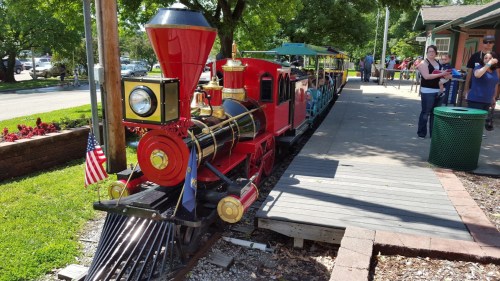 A colorful miniature train parked on a wooden platform, surrounded by trees and people enjoying the day.