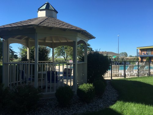 A gazebo surrounded by greenery, with a pool and lounge chairs visible in the background under a clear blue sky.