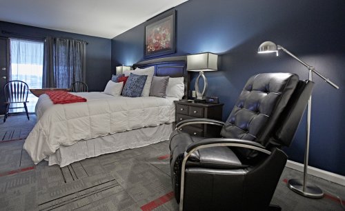 Cozy bedroom with a large bed, decorative pillows, a black chair, and a lamp against a dark blue wall.