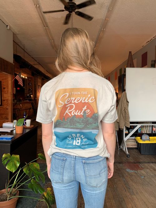A person wearing a light-colored t-shirt with a graphic design that says "Took the Scenic Route" on the back.