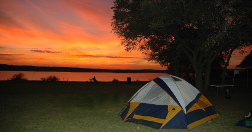 A colorful sunset over a lake with a tent set up on the grassy shore, surrounded by trees.