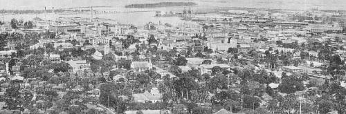 Aerial view of a historic cityscape with buildings, trees, and a waterfront in the background.