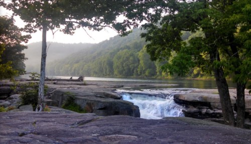 A serene river scene with a waterfall, surrounded by lush green trees and rocky terrain under a soft morning light.
