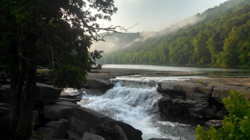 A serene river scene with a waterfall, surrounded by lush green trees and misty mountains in the background.
