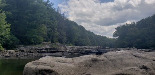 A serene river scene with rocky banks, surrounded by lush green trees under a partly cloudy sky.