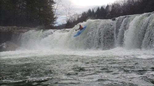 A kayaker leaps off a waterfall, splashing into the turbulent water below, surrounded by trees and mist.