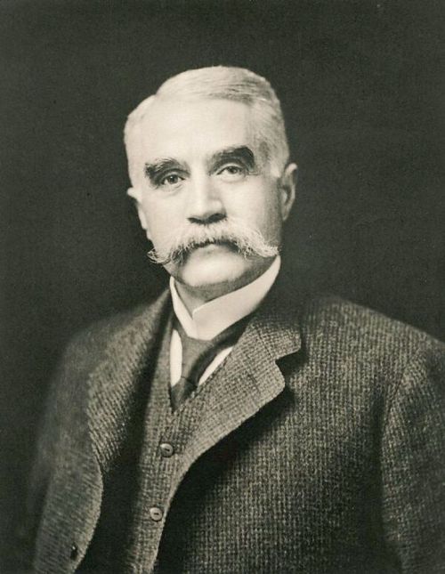 A black-and-white portrait of a man with a prominent mustache, wearing a suit and vest, looking directly at the camera.