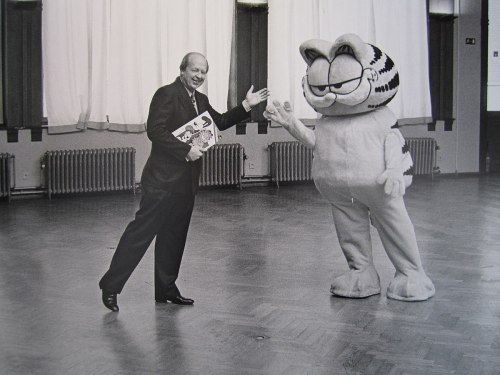 A man in a suit waves at a large Garfield mascot in a spacious, empty room with wooden floors.