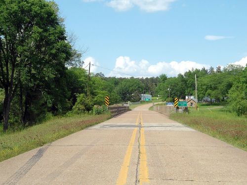 A rural road leads to a bridge, surrounded by greenery and a few buildings under a blue sky with clouds.
